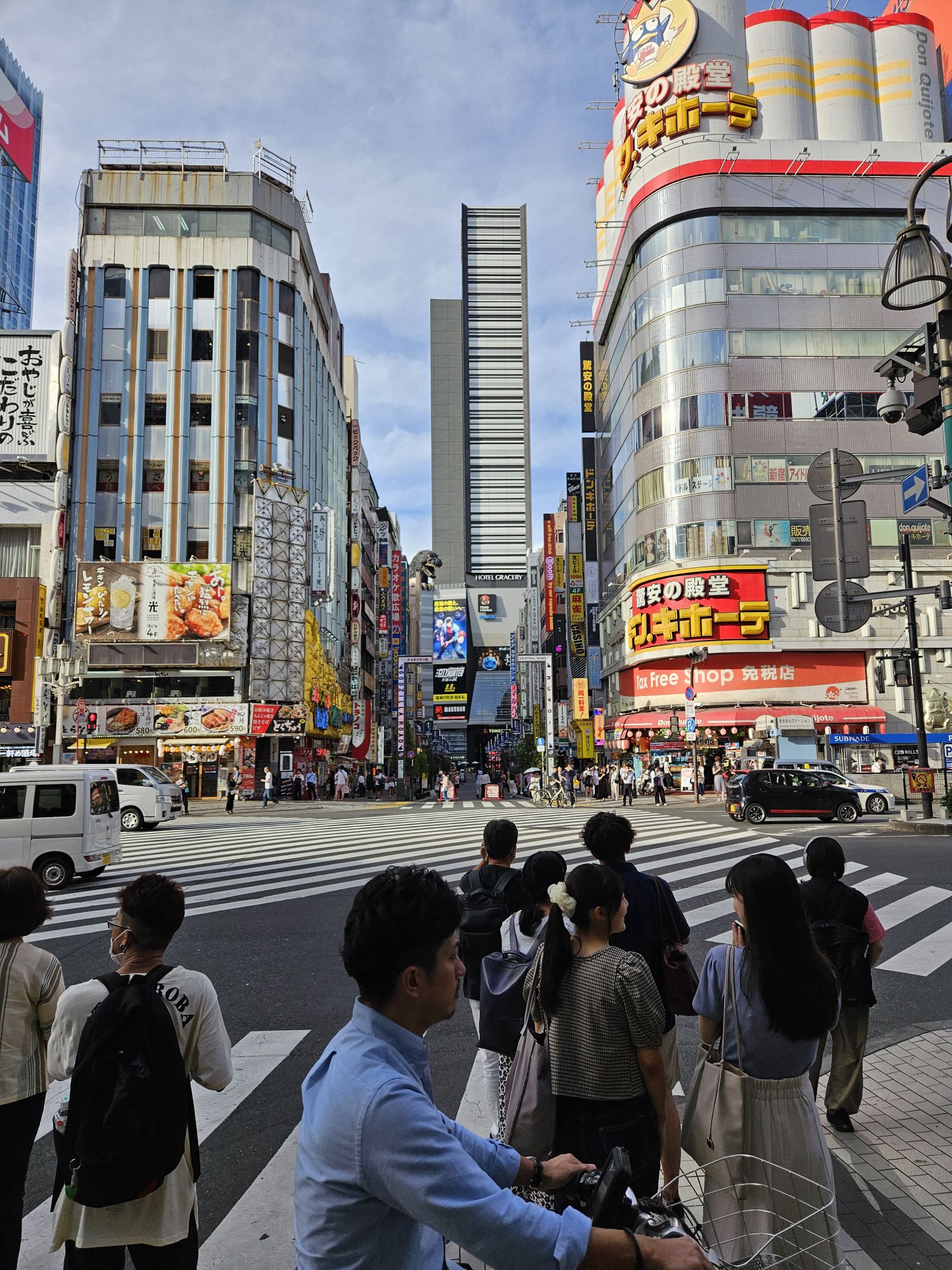 A busy Tokyo street scene with people waiting at a crosswalk. Tall buildings display colorful billboards and advertisements in Japanese. A bright blue sky with scattered clouds is visible above. A cyclist in the foreground wears a blue shirt and white pants, waiting to cross.